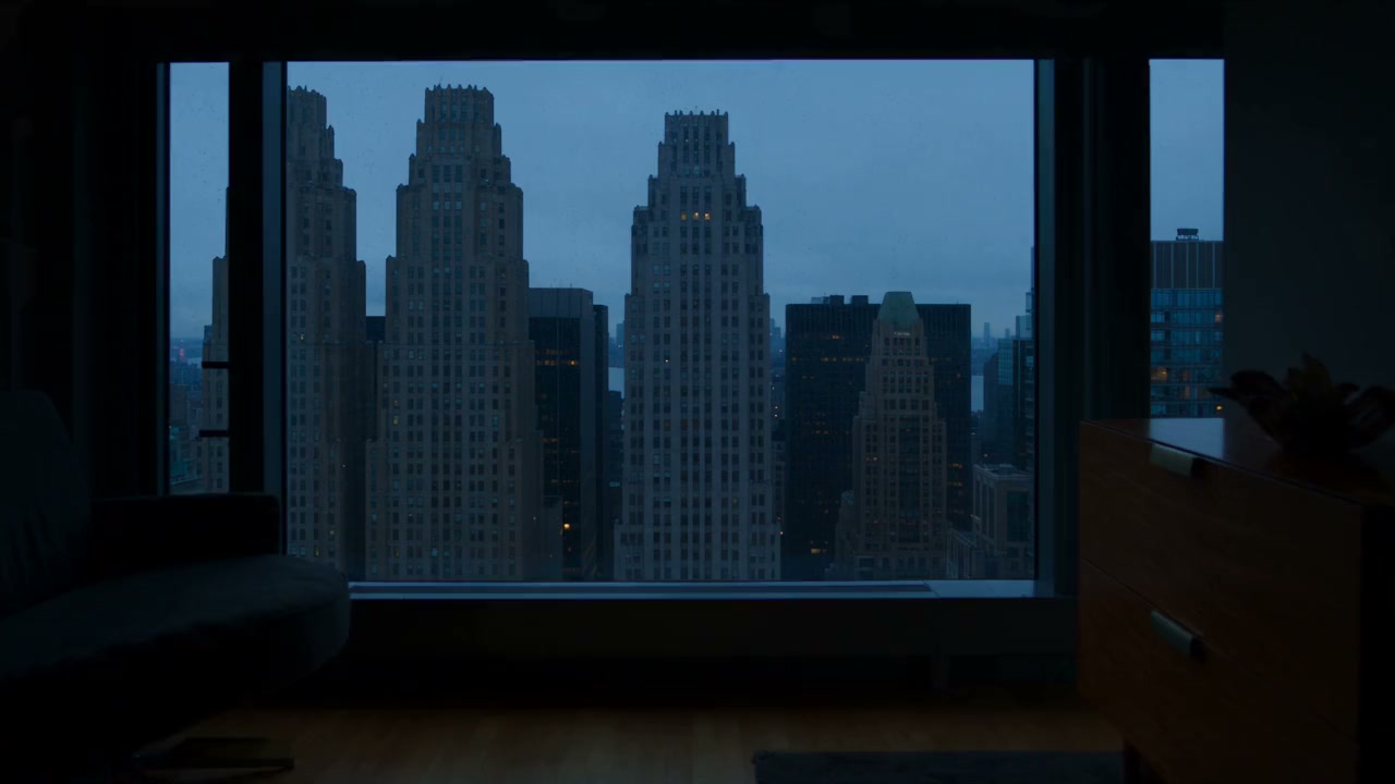A view through a wide window of stone office towers at blue hour, a few windows lit and rain spots on the glass.