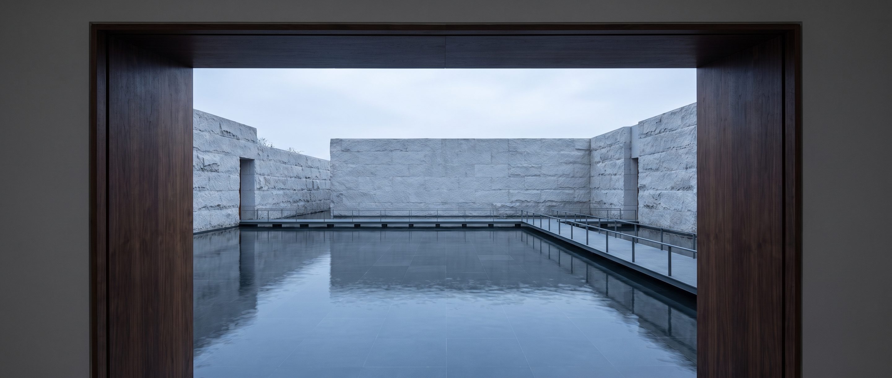 A still reflecting pool against a plain stone wall, overcast light.