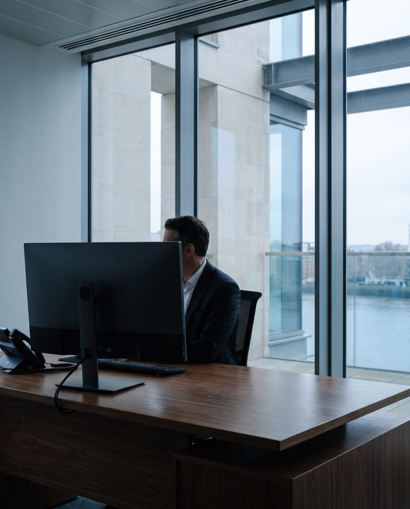 An adult in business dress at a desk, three-quarter view from behind a monitor.