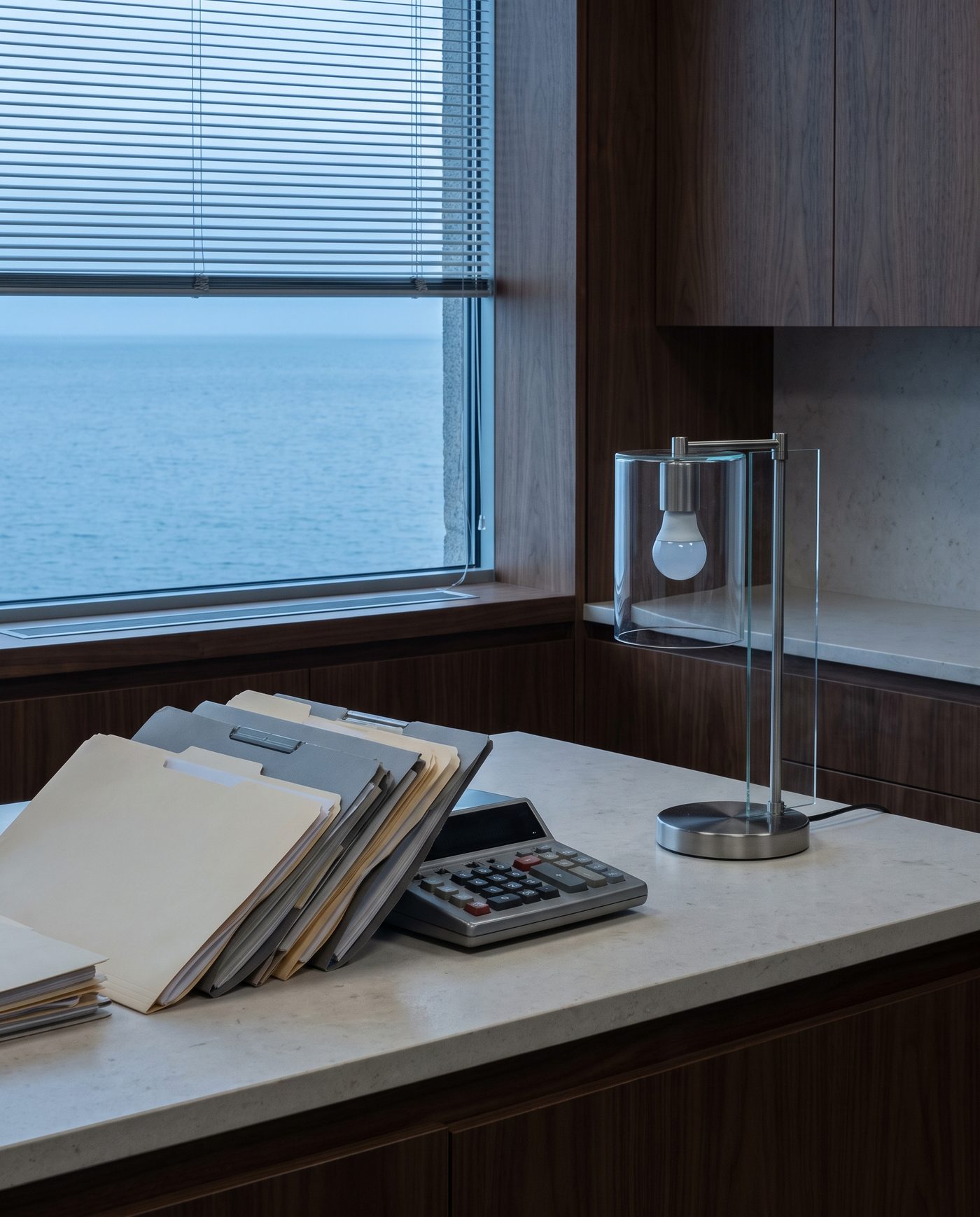 Stacked folders and a calculator beside a desk lamp on a pale stone counter.